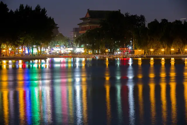 Houhai Lake at night, with coloured lights from the lakeside reflected in the water and the Drum Tower behind the trees.