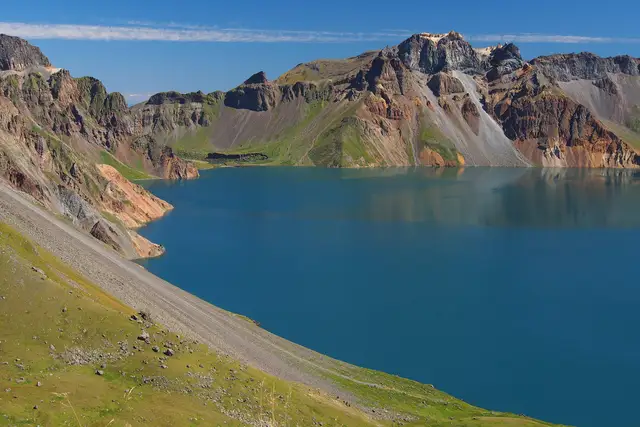The deep blue Heaven Lake inside the crater of Changbai Mountain, ringed by ochre and green volcanic peaks under a clear sky, seen from the western rim in Jilin