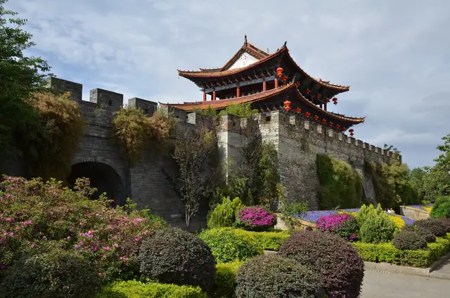 The South Gate tower of Dali old town rising above the grey stone city wall, with flowering shrubs in the garden below.
