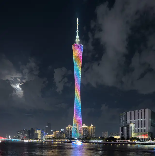 Canton Tower lit in rainbow colours beside the Pearl River in Guangzhou at night, under a cloudy sky with the moon behind the clouds.