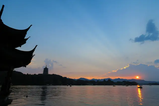 Sunset over West Lake in Hangzhou, with silhouetted pavilion eaves in the foreground, Leifeng Pagoda on the far shore and small boats on the water.