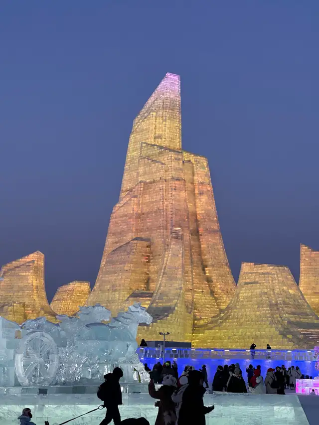 A tall illuminated ice tower and an ice sculpture of horses at Harbin Ice and Snow World at dusk, with visitors in silhouette.