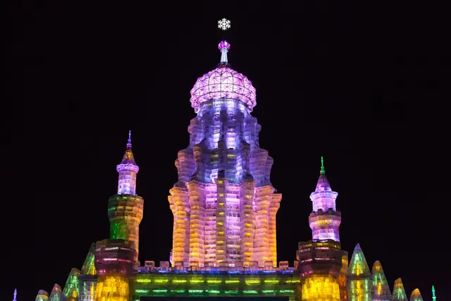 A domed tower built of ice blocks, lit purple, yellow and green against the night sky at the Harbin ice festival.