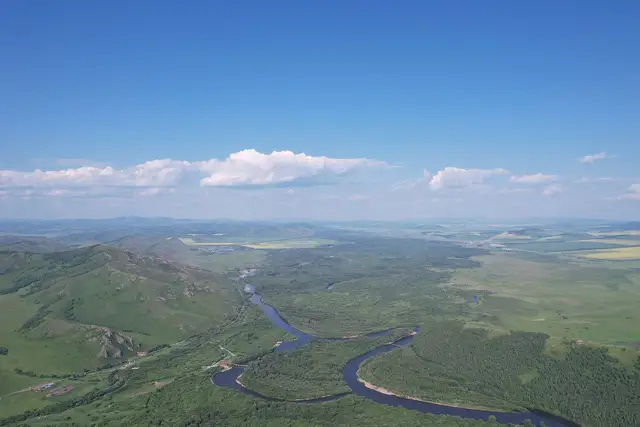 Aerial view of the Gen River winding through green grassland and forest in Ergun National Wetland Park, Hulunbuir, under a wide summer sky.