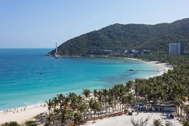 A turquoise bay in Sanya, Hainan, with a white-sand beach lined by coconut palms and a green headland under a clear sky