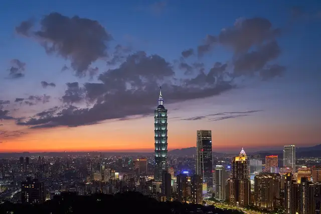 Taipei 101 lit up at dusk above the Xinyi district skyline, seen from Elephant Mountain under an orange-and-blue sunset sky with dark clouds.