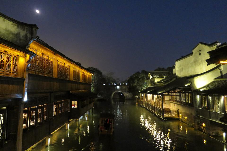 A canal in Wuzhen's West Scenic Zone at night, with lantern-lit timber houses on both banks, a small boat on the water and the moon overhead.