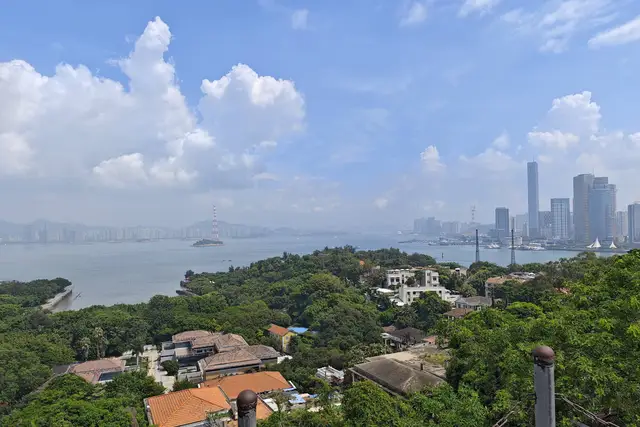 Red-tiled villas among green trees on Gulangyu Island, with the sea and the Xiamen skyline beyond under a blue sky